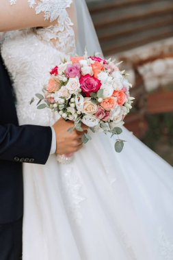 Bir gelin ve damat el ele tutuşuyor ve gelin bir buket çiçek tutuyor. Buket pembe ve beyazdır ve çiçek karışımı içerir. Sahne romantik ve mutlu.