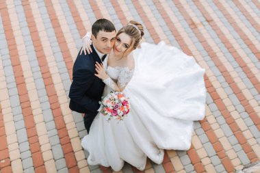 Bir gelin ve damat, tuğla kaldırımda fotoğraf çektirmek için poz veriyor. Gelin beyaz bir elbise giyiyor ve elinde bir buket çiçek tutuyor. Adam takım elbise giyiyor ve onun arkasında duruyor.