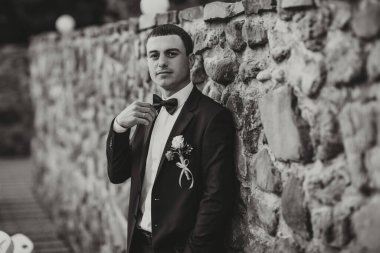 Takım elbiseli ve kravatlı bir adam taş bir duvarın önünde duruyor. Papyon takıyor ve elinde bir gül tutuyor. Zarafet ve incelik kavramı