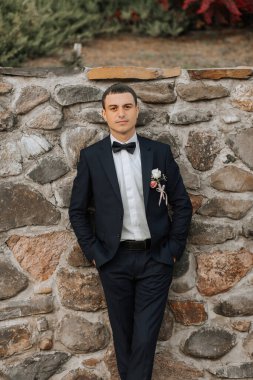 Takım elbiseli ve papyonlu bir adam taş bir duvarın önünde duruyor. Yakasına yaka çiçeği takmış.