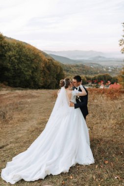 Gelin ve damat, gelin beyaz bir elbise ve damat takım elbise giyerek bir tarlada duruyorlar. Birbirlerini kucaklıyorlar ve gelin elinde bir buket tutuyor. Huzur dolu bir sahne.