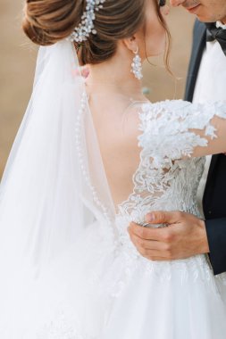 Bir gelin ve damat düğün fotoğrafında birbirlerine sarılıyorlar. Gelin duvağı ve çiçekli başlığı olan beyaz bir elbise giyiyor. Fotoğraf aşkı yakalar.