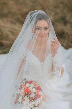 Bir gelin kucağında bir buket çiçekle yerde oturuyor. Duvak ve beyaz bir elbise giyiyor.