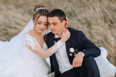 Bir gelin ve damat çimlerin üzerinde oturuyor, birbirlerine sarılıyorlar. Gelin beyaz bir elbise giyiyor ve damat bir takım elbise giyiyor. Sahne romantik ve samimi.