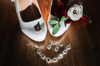 Ayak parmağında gümüş bir tılsım ve topuğunda kırmızı bir gül olan bir çift beyaz ayakkabı. Masada gümüş bir bilezik ve bir çift küpe de var.