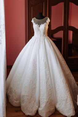 Üzerinde bir sürü parıltı olan uzun beyaz bir elbise. Elbise pembe duvarlı bir odada asılı.