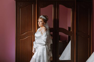 Beyaz elbiseli bir kadın ahşap bir dolabın önünde duruyor. Bir taç ve duvak takıyor.