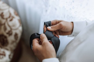 Bir adam kravatını bağlıyor. Kravat siyah ve adam beyaz gömlek giyiyor.