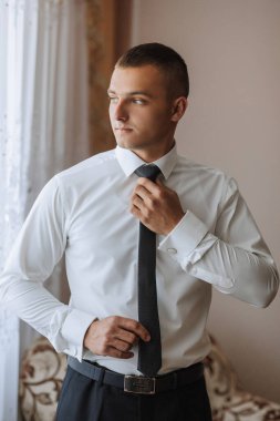 Beyaz gömlekli ve siyah kravatlı bir adam pencerenin önünde duruyor. Kravatını düzeltiyor ve pencereden dışarı bakıyor. Adam takım elbise giydiği için resmiyet ve profesyonellik kavramı.