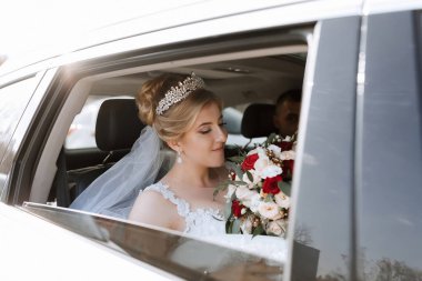 Bir gelin elinde bir buket çiçekle arabada oturuyor. Bir taç ve duvak takıyor.