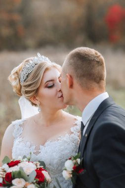 Kızıl bir çalılığın önünde bir gelin ve damat öpüşüyor. Gelin taç takıyor ve damat da takım elbise giyiyor.