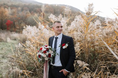 Takım elbiseli ve kravatlı bir adam elinde bir buket çiçekle uzun otların arasında duruyor.