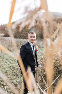 Takım elbiseli ve kravatlı bir adam uzun otların arasında duruyor. Yakasında kırmızı bir gül var.