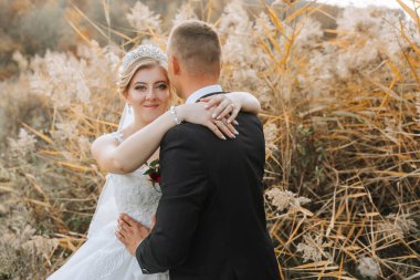 Bir gelin ve damat uzun otların arasında kucaklaşıyor. Damat takım elbise giyerken gelin beyaz bir elbise ve taç giyiyor. Sahne samimi ve romantik, aşkı yakalıyor.