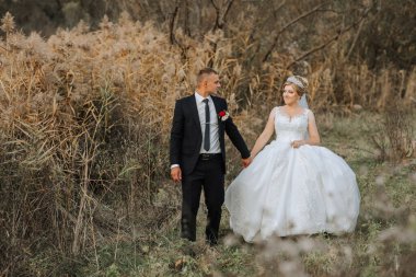 Bir gelin ve damat uzun otların arasında yürüyorlar. Gelin beyaz bir elbise giyiyor ve damat bir takım elbise giyiyor. Sahne romantik ve huzurlu.