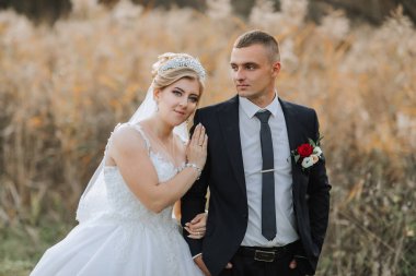 Gelin ve damat, gelin beyaz bir elbise ve damat takım elbise giyerek bir tarlada dururlar.