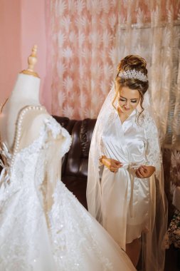 Gelinin biri gelinliğin önünde durmuş, ona bakıyor. Elbise beyaz ve uzun bir treni var. Gelin beyaz bir duvak ve taç takıyor. Sahne kanepeli bir odada geçiyor.