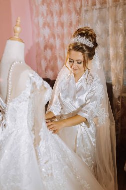 Bir gelin düğünü için hazırlanıyor ve bir elbisenin önünde duruyor. Bir taç ve duvak takıyor.
