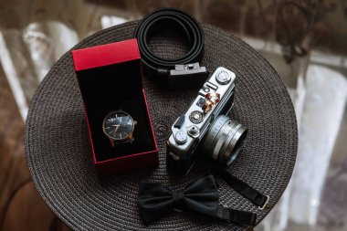 Masada kırmızı bir kutu ve siyah bir papyonun yanında gümüş bir kamera duruyor. Kamera bir saat ve siyah kuşak da dahil olmak üzere diğer nesnelerle çevrilidir.