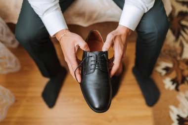 Bir adam elinde siyah bir ayakkabı tutuyor. Ayakkabı siyah ve üzerinde dantel var. Adam bir yatakta oturuyor.