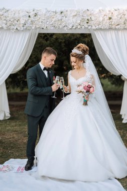 Gelin ve damat bir gölgeliğin altında duruyorlar, gelin bir bardak şampanya tutuyor. Gelin beyaz bir elbise giyiyor ve damat da takım elbise giyiyor.