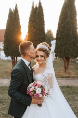 Bir gelin ve damat bir ağacın önünde birbirlerinin yanaklarını öpüyor. Gelin beyaz bir elbise giyiyor ve damat yeşil bir takım giyiyor.