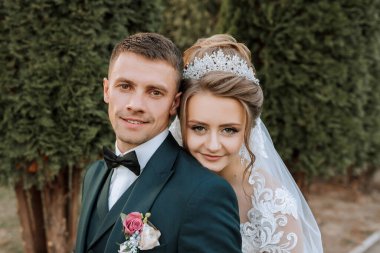 Bir gelin ve damat bir ağacın önünde fotoğraf için poz veriyorlar. Gelin peçe takıyor ve damat takım elbise giyiyor.