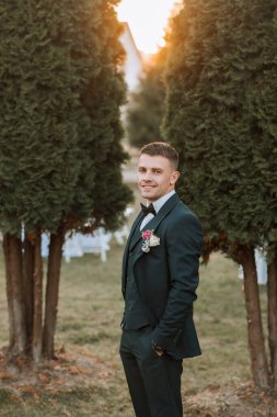 Takım elbiseli ve kravatlı bir adam iki ağacın önünde duruyor. Gülümsüyor ve yakasına kırmızı bir çiçek takıyor.