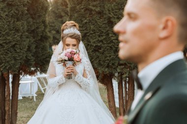 Bir gelin gelinlik giyiyor ve elinde bir buket çiçek tutuyor. Arkasında bir damat duruyor, ona bakıyor.