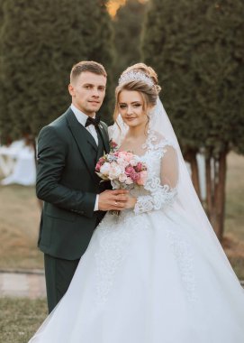 Bir gelin ve damat bir ağacın önünde fotoğraf için poz veriyorlar. Gelin beyaz bir elbise giyiyor ve damat bir takım elbise giyiyor. Gelin elinde bir buket çiçek tutuyor.