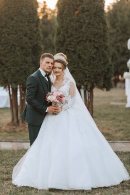 Bir gelin ve damat bir ağacın önünde fotoğraf için poz veriyorlar. Gelin uzun beyaz bir elbise giyiyor ve damat bir takım elbise giyiyor. Çift ellerinde çiçekleri tutuyor.