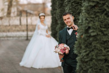 Bir gelin ve damat bir çitin önünde duruyor. Gelin beyaz bir elbise giyiyor ve damat bir takım elbise giyiyor. Damat elinde bir buket çiçek tutuyor.