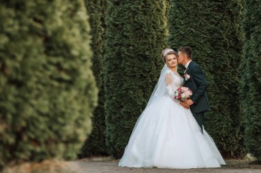 Bir gelin ve damat çitin önünde fotoğraf için poz veriyorlar. Gelin beyaz bir elbise giyiyor ve damat bir takım elbise giyiyor. İkisi de gülümsüyor ve kameraya bakıyor.