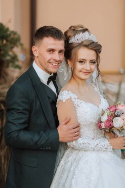 Bir gelin ve damat fotoğraf için poz veriyor. Gelin bir gelinlik ve taç giyiyor. Çift elinde bir buket çiçek tutuyor.