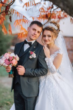 Bir gelin ve damat bir ağacın önünde fotoğraf için poz veriyorlar. Gelin beyaz bir elbise giyiyor ve damat bir takım elbise giyiyor. Gelin bir buket tutuyor.