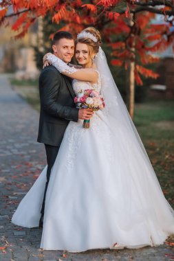 Bir gelin ve damat bir ağacın önünde fotoğraf için poz veriyorlar. Gelin beyaz bir elbise giyiyor ve damat bir takım elbise giyiyor. Ellerinde çiçekler tutuyorlar.