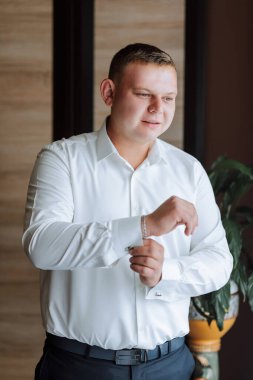 Beyaz gömlekli bir adam giyiniyor ve kelepçesini ayarlıyor. Formalite anlayışı ve detaylara dikkat, adam görünüşünün düzgün ve parlak olduğundan emin olmak için özen gösteriyor.