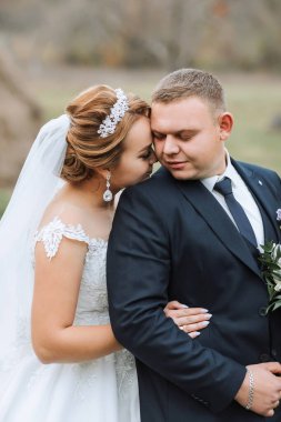 Bir gelin ve damat düğün fotoğrafında birbirlerine sarılıyorlar. Gelin beyaz bir elbise ve taç giyerken damat takım elbise ve kravat takıyor. Fotoğraf bir aşk anını yakalar.