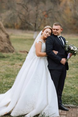 Bir gelin ve damat tarlada fotoğraf çektirmek için poz veriyorlar. Gelin beyaz bir elbise giyiyor ve damat da takım elbise giyiyor.