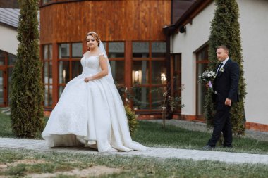 Gelin ve damat bir evin önünde duruyorlar, gelin beyaz bir elbise giyiyor ve damat takım elbise giyiyor.