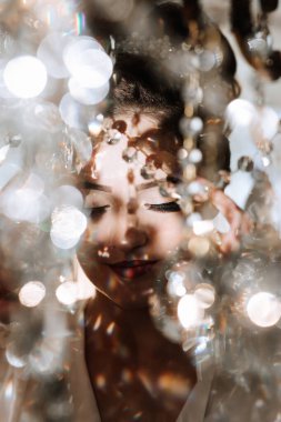 Bir kadın yüzünde bir gülümsemeyle kameraya bakıyor. Etrafında bir sürü parıltı var, bu da görüntüye ışıl ışıl ve neşeli bir hava katıyor.