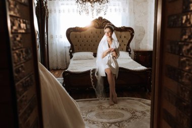 Gelinlik giymiş bir kadın, odadaki bir yatakta oturur. O bir peçe takıyor ve bir çanta tutuyor.