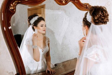 Bir kadın düğünü için hazırlanıyor ve aynada kendine bakıyor. Beyaz bir elbise ve duvak takıyor.