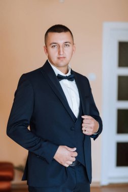 Takım elbiseli ve kravatlı bir adam beyaz bir kapının önünde duruyor. Mavi bir takım elbise ve mavi papyonlu beyaz bir gömlek giyiyor. Kravatını düzeltiyor ve kameraya bakıyor.