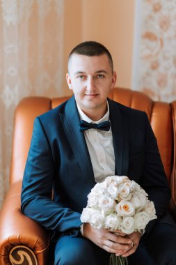 Takım elbiseli ve kravatlı bir adam kahverengi bir sandalyede oturuyor elinde bir buket beyaz çiçekle