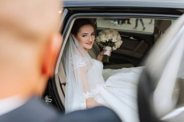 Bir gelin elinde bir buket çiçekle arabada oturuyor. Arkasında bir adam duruyor.