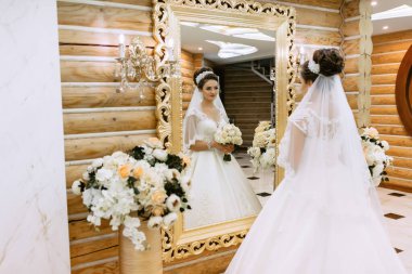 Bir gelin aynanın önünde beyaz bir elbise ve altın bir saç bandı takıyor. Elinde bir buket çiçek ve bir vazo çiçek tutuyor.