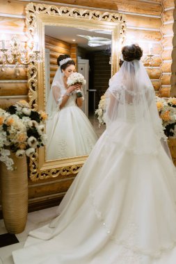 Bir gelin aynanın önünde elinde bir buket çiçekle duruyor. Ayna, gelinin güzelliğini ve zarafetini yansıtan gösterişli ve altındır. Romantizm ve kutlama kavramı