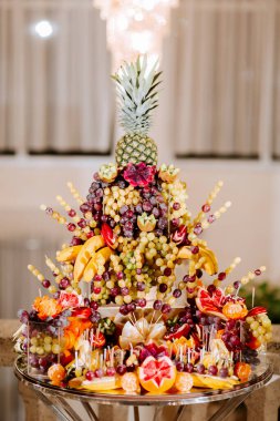 Üstünde ananas olan bir meyve sergisi. Meyve, muz, portakal ve üzümlerle bir piramit şeklinde düzenlenir. Sunum renkli ve davetkar.