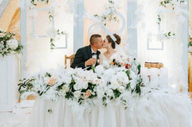 Düğün resepsiyonunda bir gelin ve damat öpüşüyor. Gelin beyaz bir elbise giyiyor ve damat siyah bir takım elbise giyiyor. Önlerindeki masa çiçeklerle kaplı.